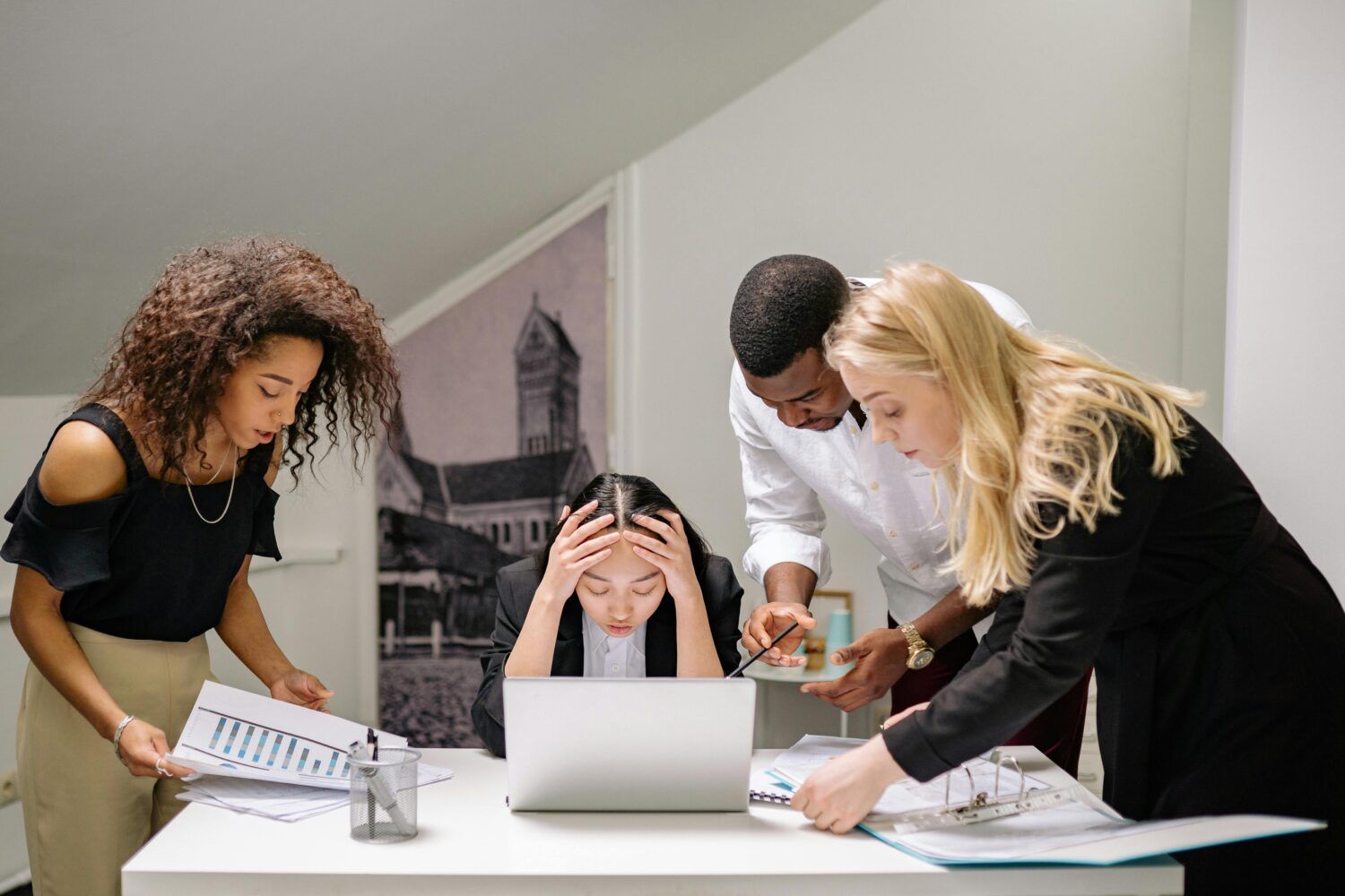 Group collaborating around a laptop in a bright office, one person stressed with head in hands