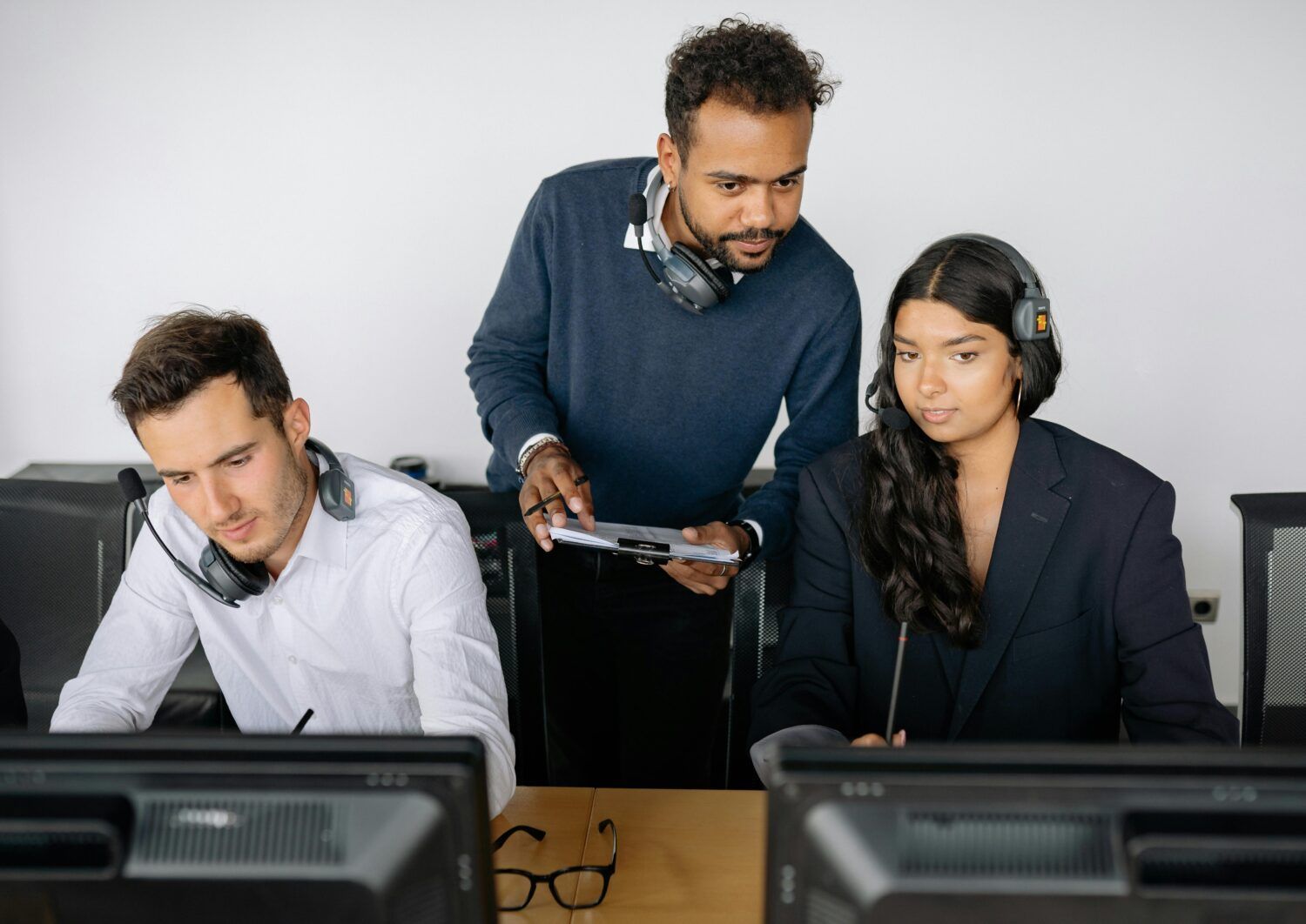Three coworkers reviewing data at office computers, with one standing and pointing at a tablet.