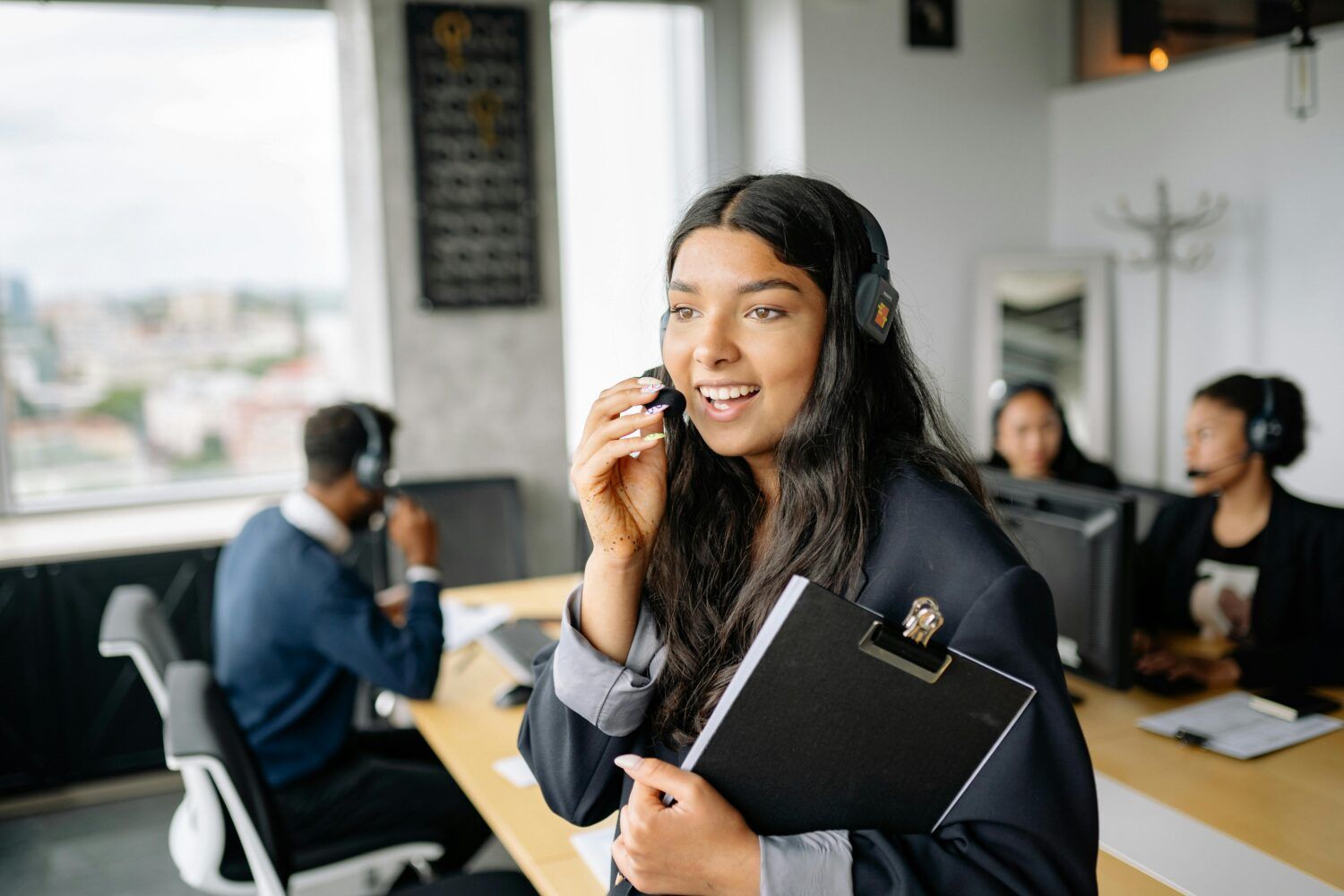 Woman wearing headphones and holding a clipboard in a modern office call center with coworkers in the background