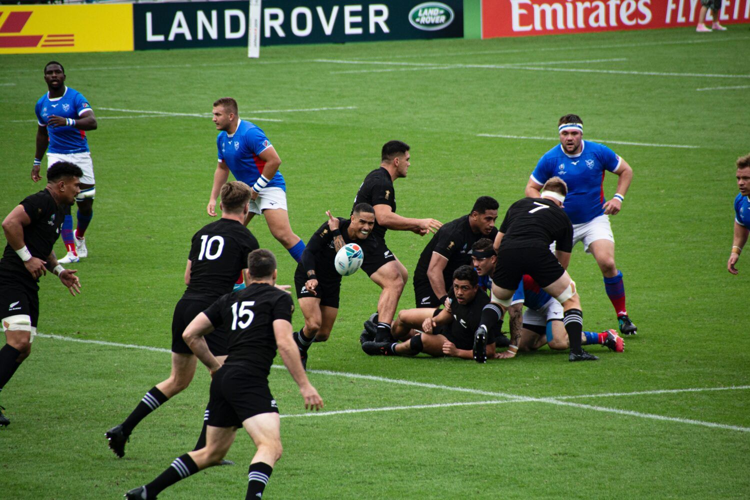 Rugby scrum near the try line, black and blue teams battling on a wet field with sponsor boards behind.