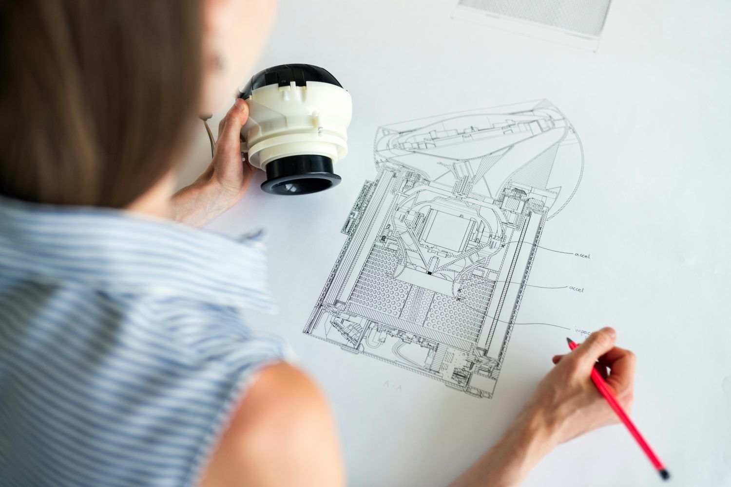 Person studying a technical drawing with a magnifier and red pencil at a white desk