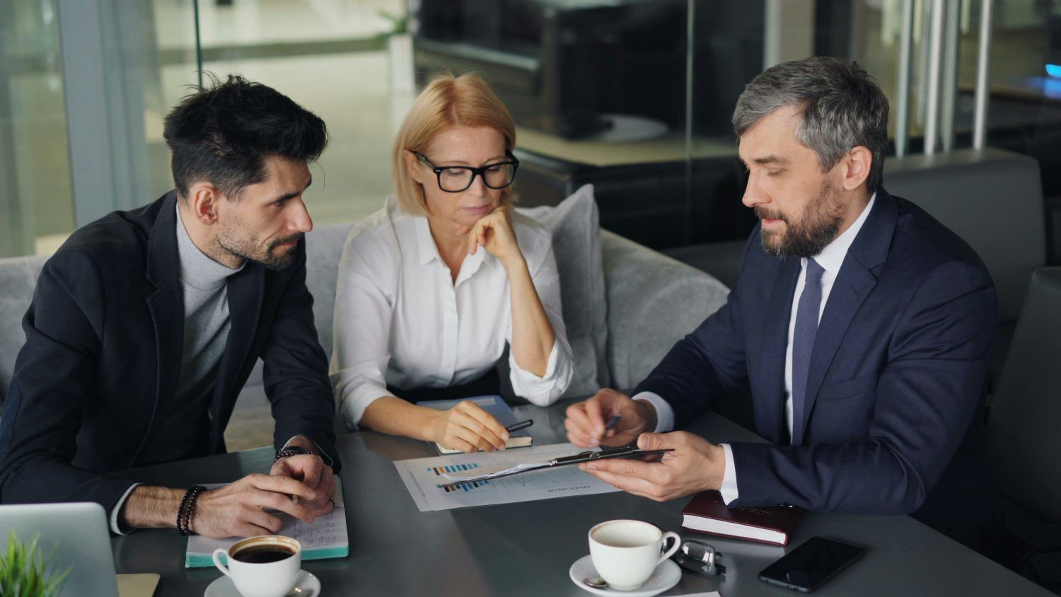 Three professionals in suits discuss documents at a conference table in a modern office.