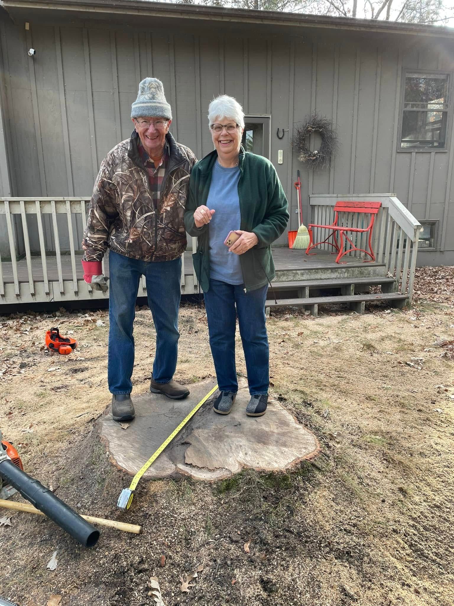 A man and a woman are standing next to a tree stump in front of a house.