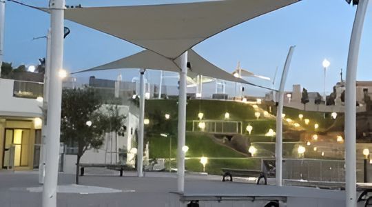 Scena di parco con vele ombreggianti, panchine e aree verdi. Pali bianchi sostengono le vele, illuminazione serale.