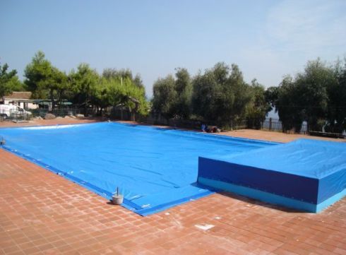 Copertura blu per piscina rettangolare su un patio in mattoni, con alberi sullo sfondo.