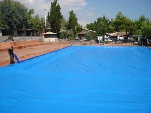 Persona che copre una piscina esterna vuota con un grande telo blu. Sullo sfondo si vedono alberi ed edifici.