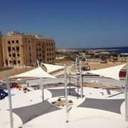 Vele ombreggianti su un'area aperta, con edifici e acqua sullo sfondo. Cielo luminoso.