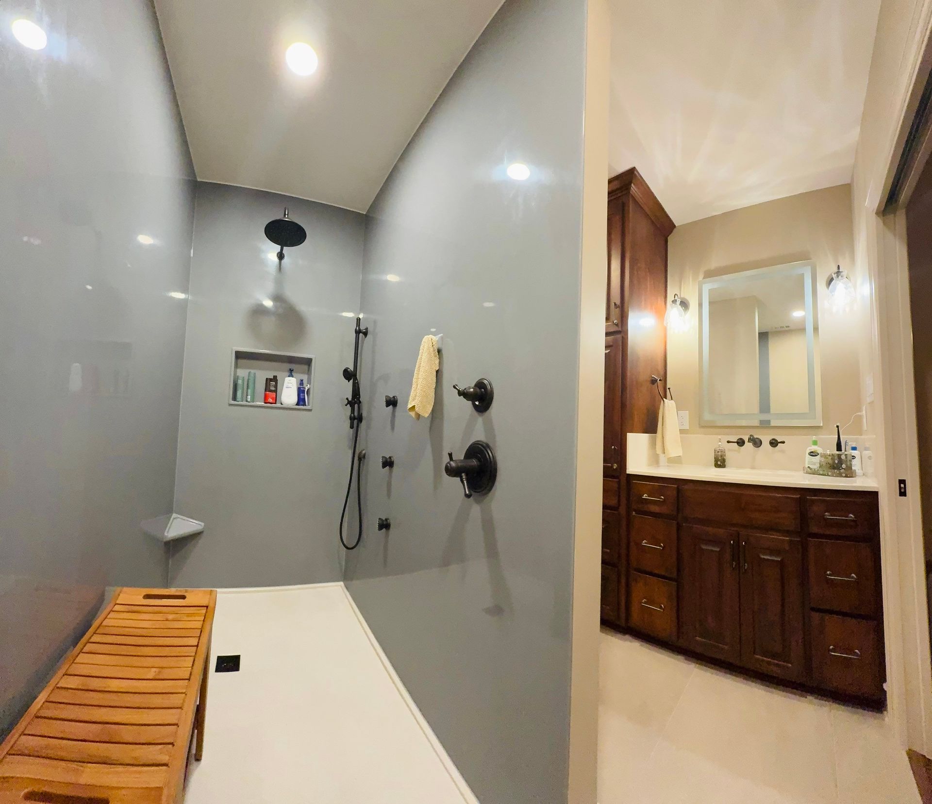 A modern bathroom featuring a walk-in shower with grey walls, a wooden bench, and a separate vanity with wood cabinets.