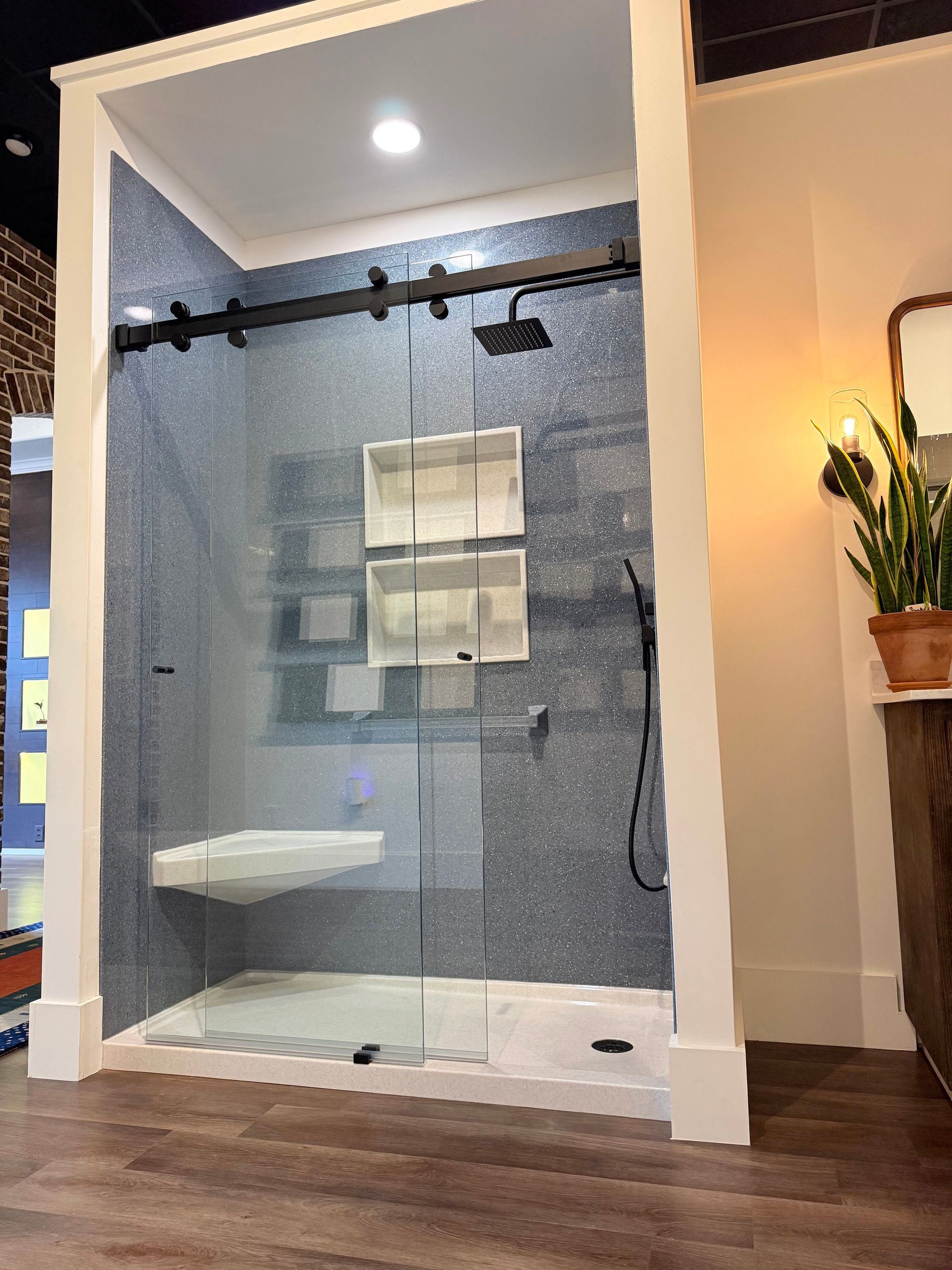 Modern shower with blue-speckled tile, glass door, white seat and shelves, dark hardware, and wooden floor.