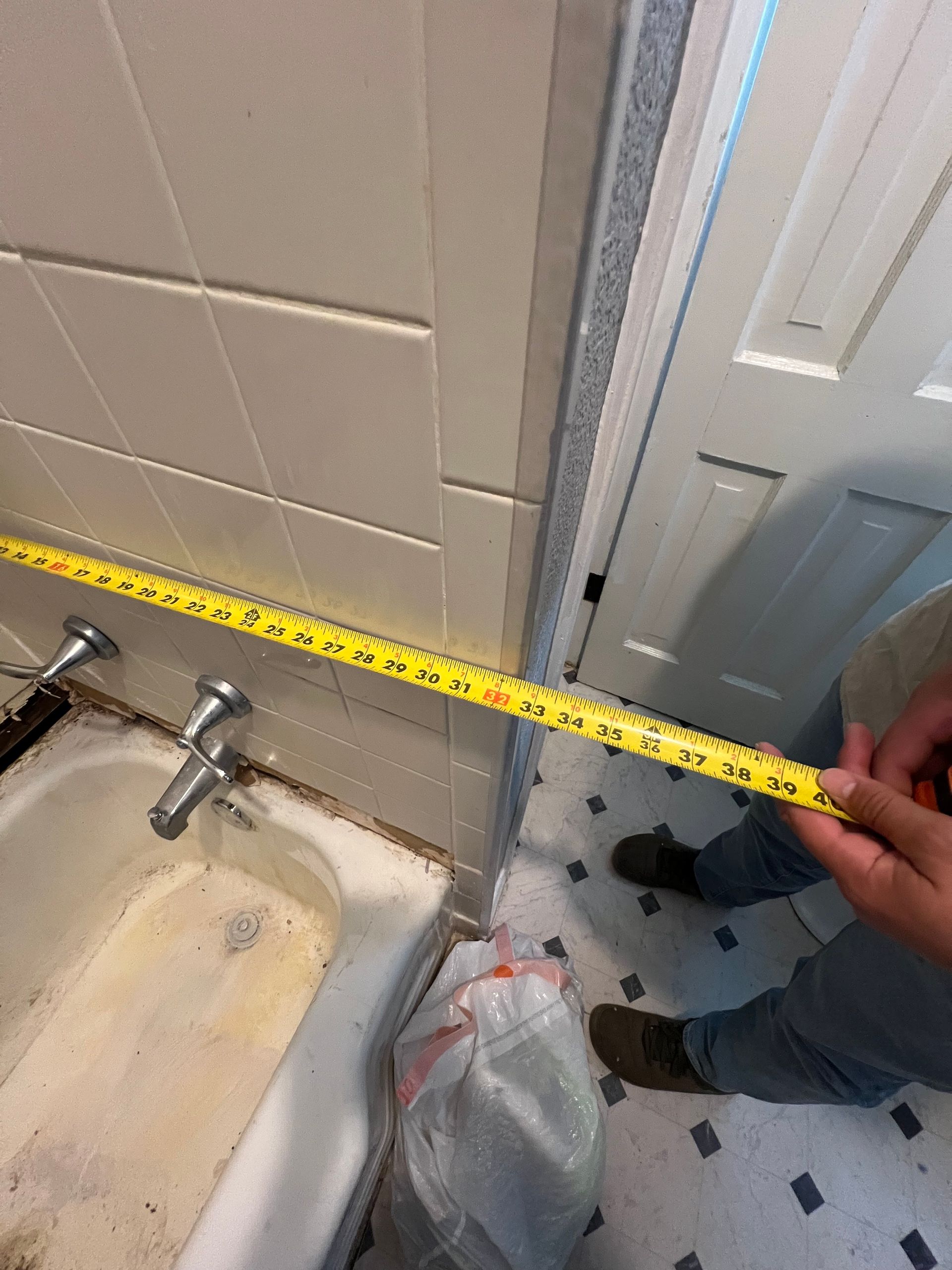 Person measuring a bathroom with a yellow tape measure, near a tub and doorway.