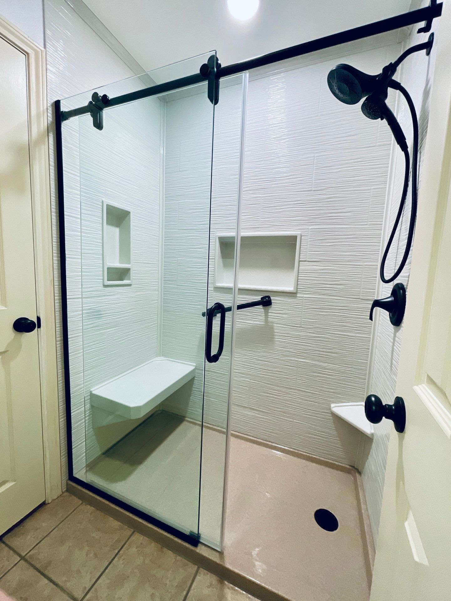 Modern bathroom shower with glass door, black fixtures, white textured tile, and built-in bench.