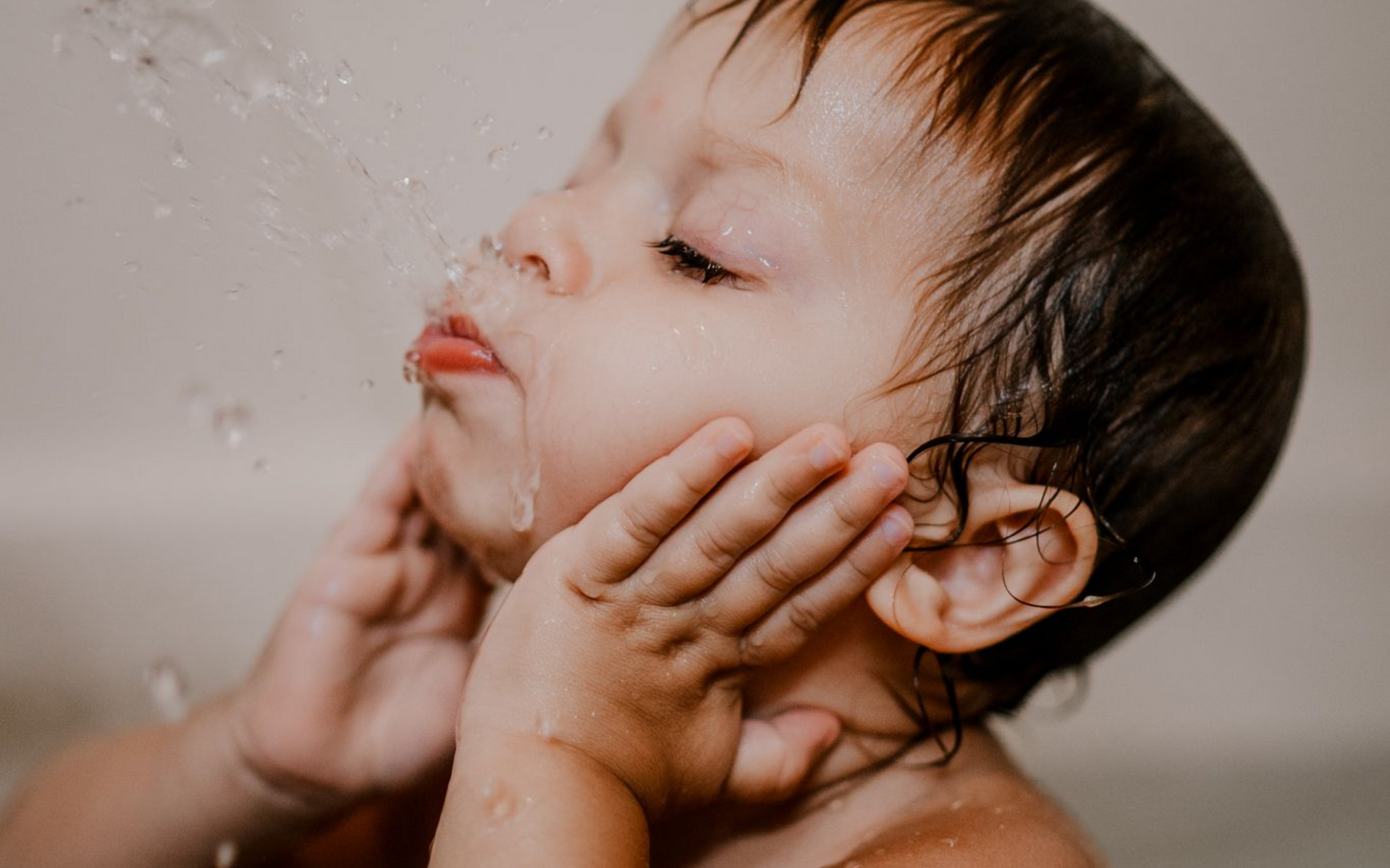 Child in a bathtub, eyes closed, getting water splashed on face. Hands on cheeks, water droplets.