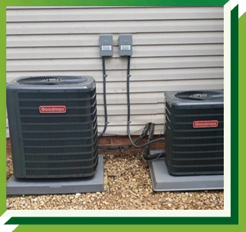 Two gray Goodman HVAC units sit on separate concrete pads against a beige siding wall with two electrical disconnect boxes.