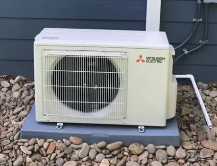 A beige Mitsubishi Electric outdoor heat pump unit sits on a grey plastic pad atop a bed of rocks against a blue wall.