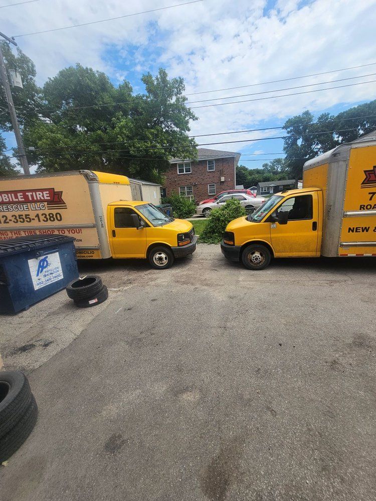 Two yellow box trucks facing each other on a paved area, with a blue dumpster and tires nearby.