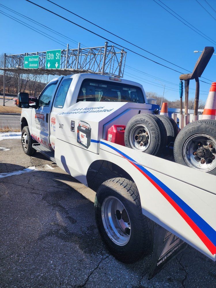 White tow truck on a road, carrying spare tires. Overhead highway sign visible. Sunny day.
