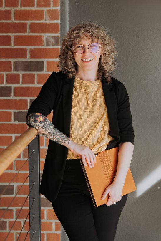 Woman with curly hair, glasses, and arm tattoos, smiling, holding a notebook, leaning against a railing, brick wall background.
