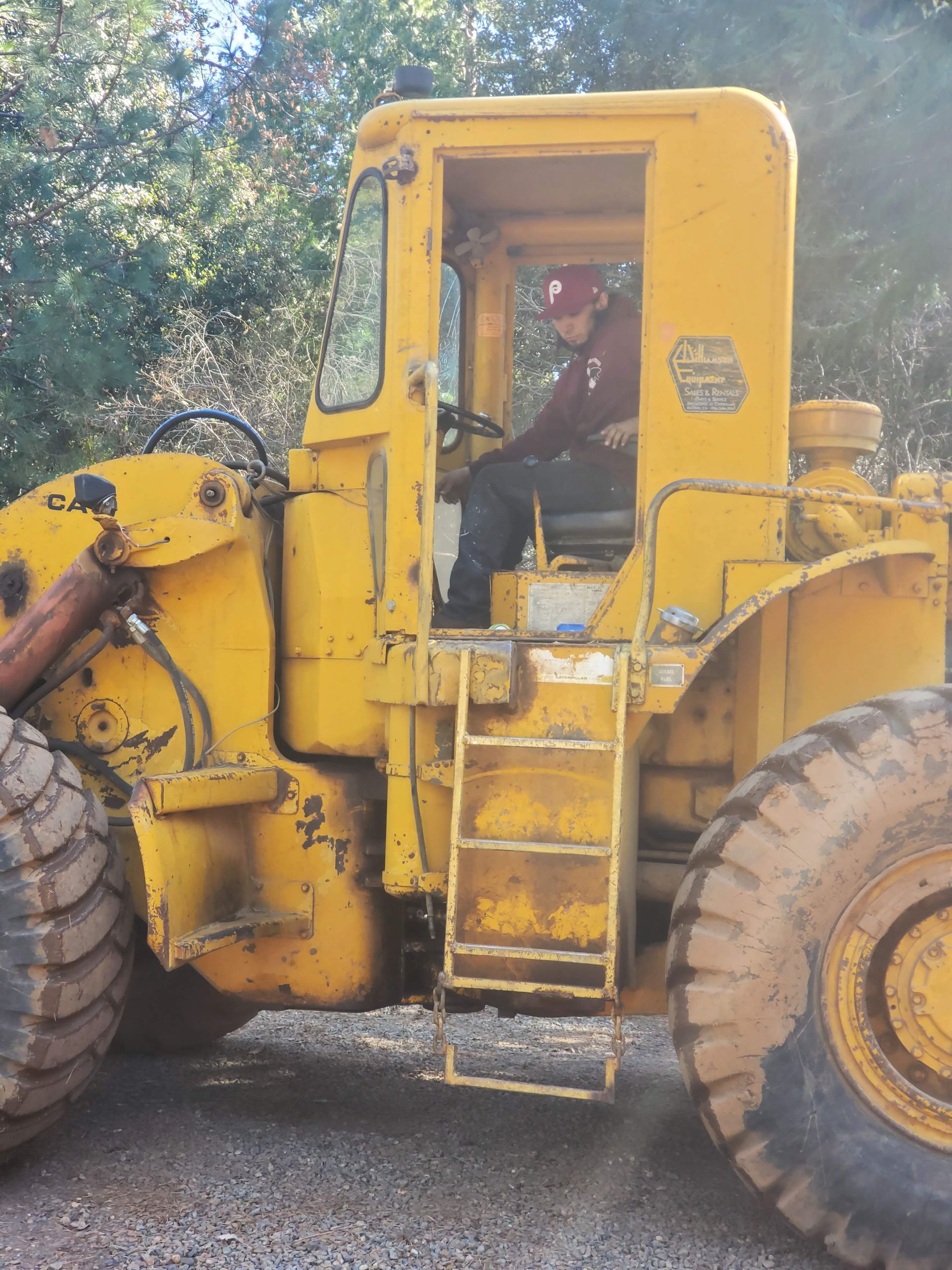 A person in a red cap sits in the driver's seat of a large yellow construction vehicle parked outdoors.