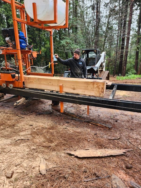 A person operates a portable orange sawmill, cutting a large wooden beam outdoors in a forest clearing.