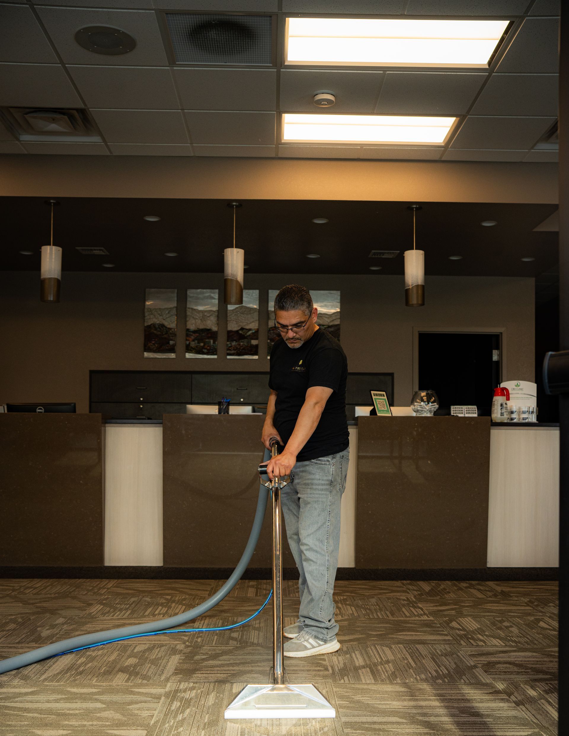 Man vacuuming carpet in an office. He's wearing jeans, a black shirt, and looking at the camera.