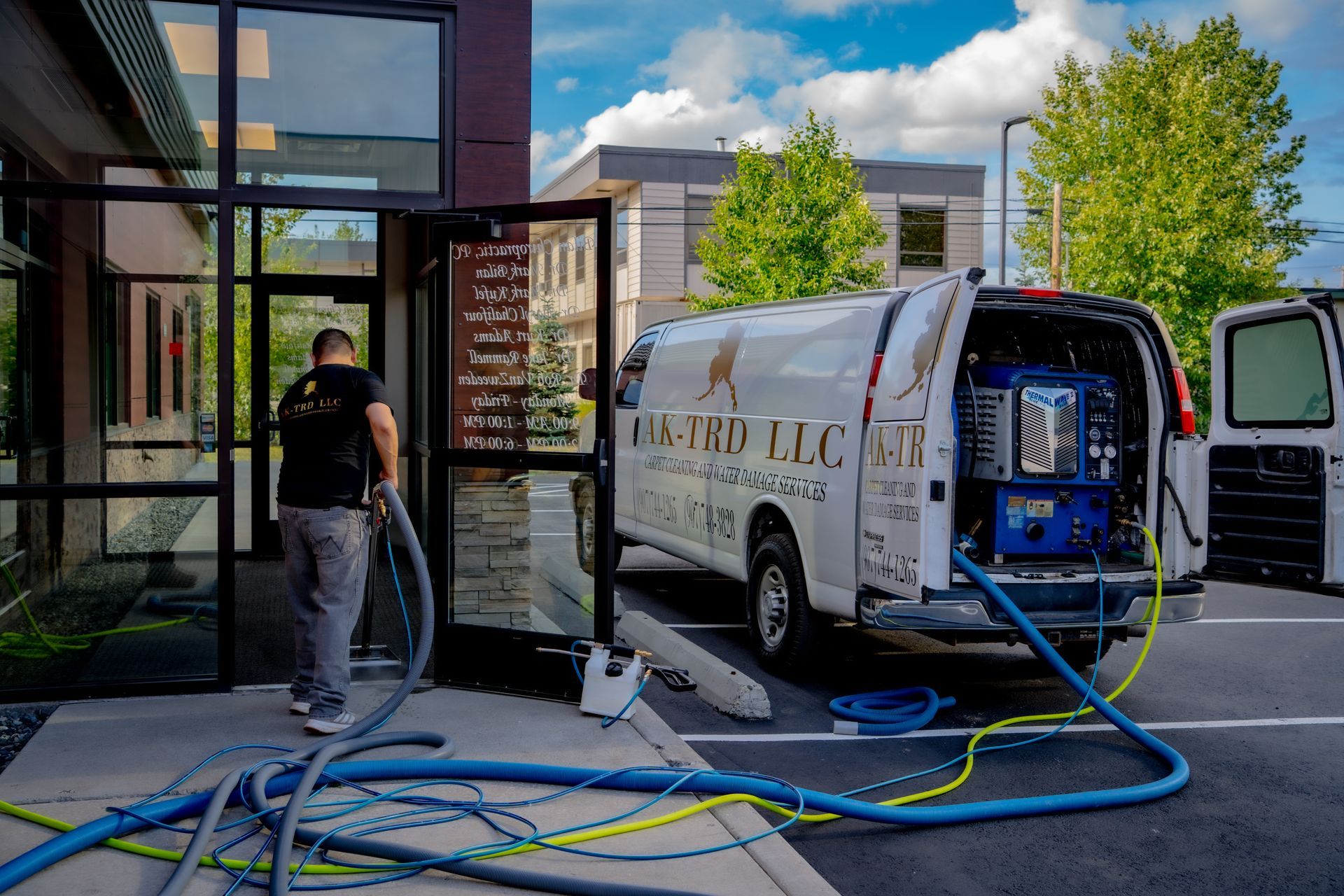 Man vacuuming a commercial building's entrance; service van with open doors and hoses visible.