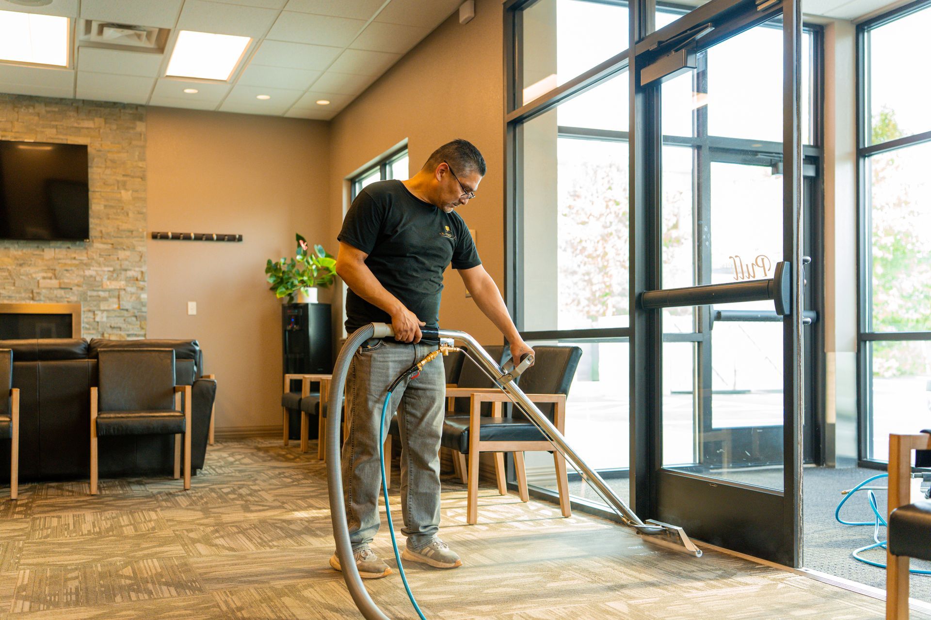 Man cleans carpet in a waiting room with a commercial carpet cleaner, near a glass door.