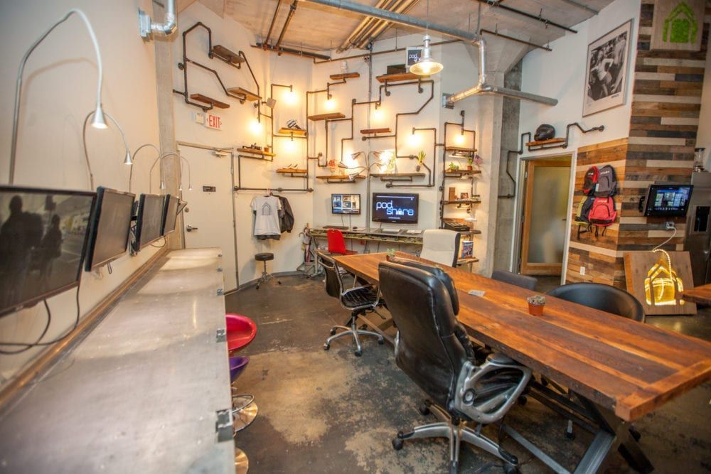 Industrial-style office space. Long wood table with black chairs. Exposed pipes and light fixtures. Monitors on a desk.