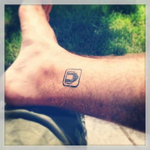 Tattoo of a phone with an arrow on a person's ankle, outdoors on a sunny day.