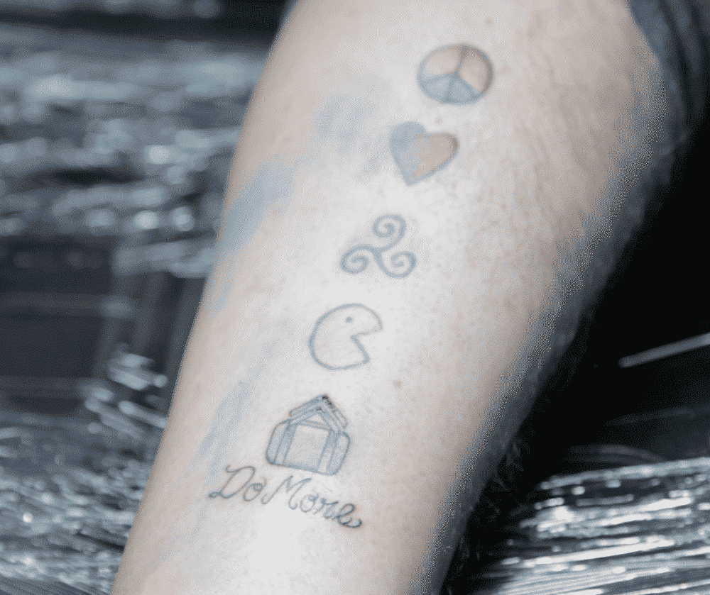 Arm with several small tattoos, including a peace sign, heart, swirl, Pac-Man, and a house; 