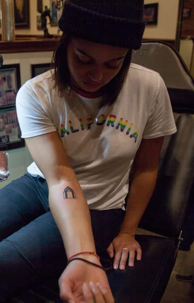 Woman with beanie and California shirt, showing a small arch tattoo on her forearm.