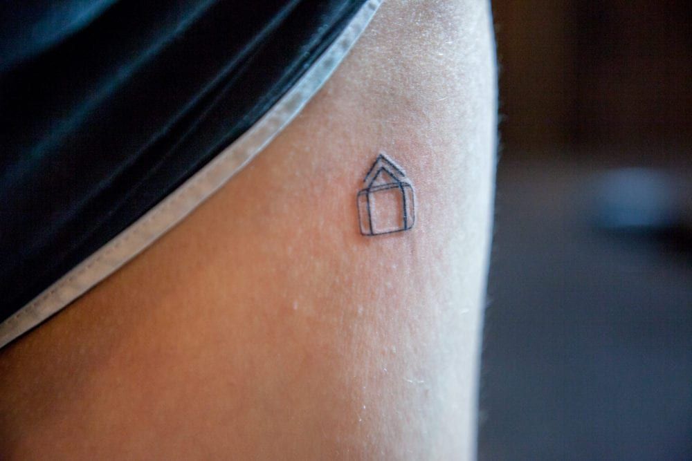 Small, simple house tattoo on a person's thigh, near the hem of black athletic shorts.