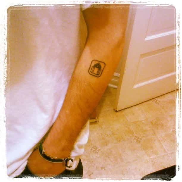 Arm with a small, framed tattoo of an arched doorway; person's leg with black shoe visible; interior setting.