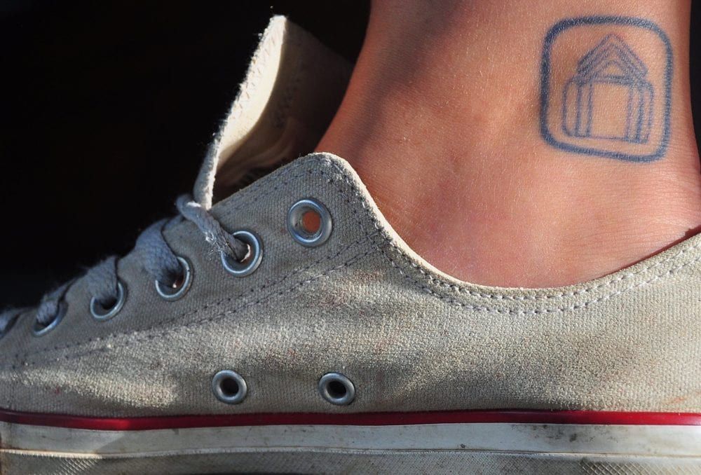 A person's ankle with a blue tattoo, next to a white Converse sneaker.