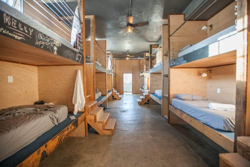 Dorm room with wooden bunk beds lining the walls; stairs lead to each bed; room has concrete floors, and light from door.