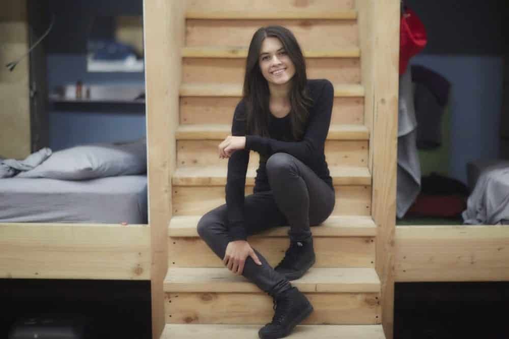 Woman sits on wooden stairs between bunk beds; she wears black and smiles.