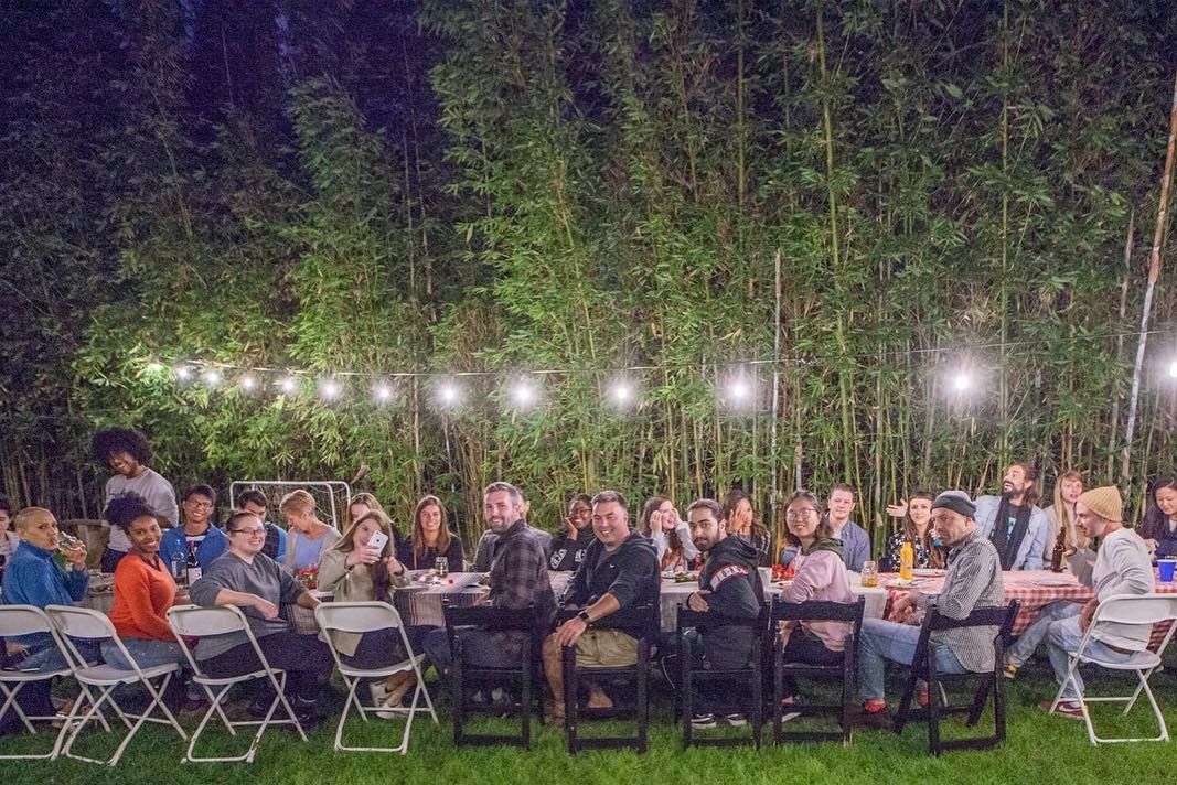 People seated at tables outdoors under string lights, with bamboo backdrop.