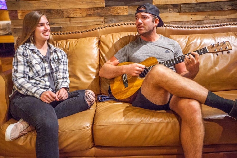 Woman smiles at man playing guitar on brown couch, rustic wood background.