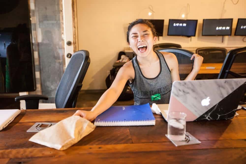 Woman with mouth open, excited, at a desk.  Laptop, notebooks, and paper bag. Office setting with screens.