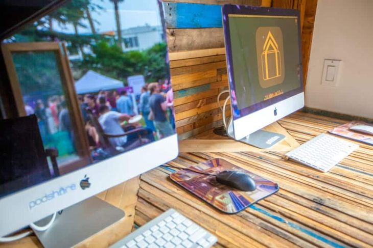 Two Apple computer monitors on a wooden desk. One displays a crowd, the other a gold-colored arrow icon.