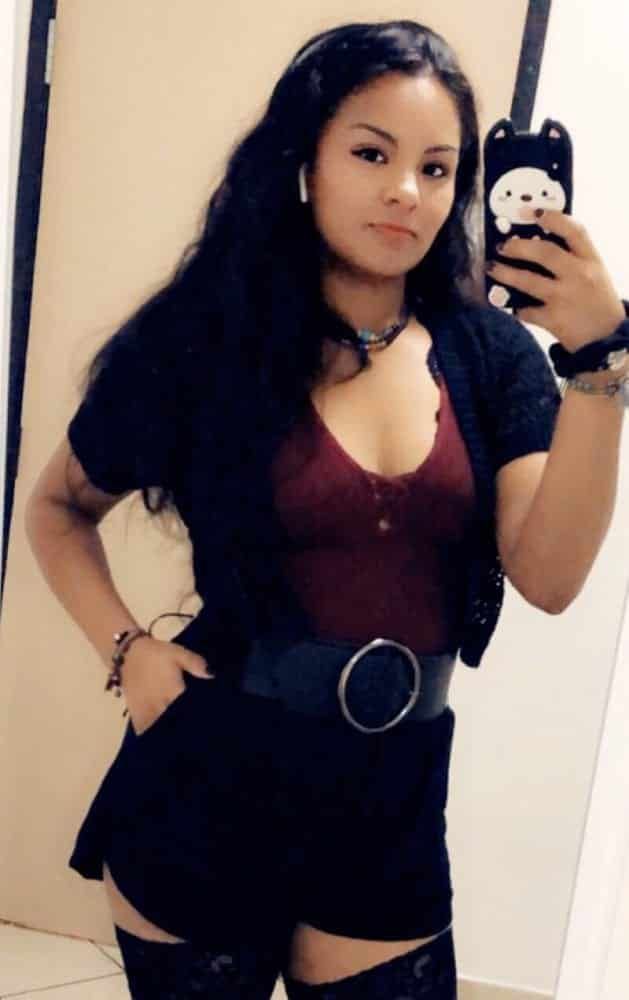 Woman taking a mirror selfie. She wears black shorts, a dark red top, a black sweater, and a wide belt.