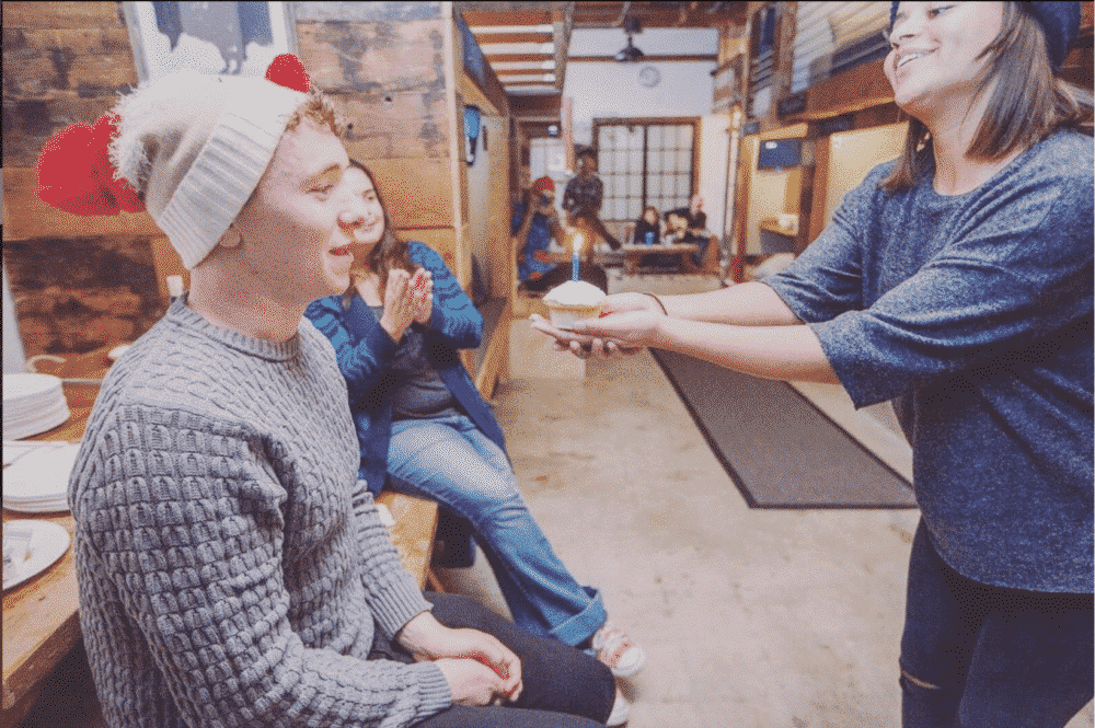 A person with a white hat and sweater, holding a cupcake with a lit candle,