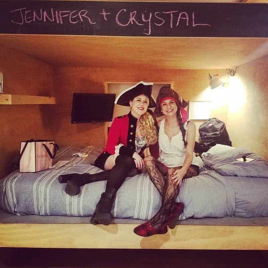 Two women in pirate costumes sitting on a bed. 