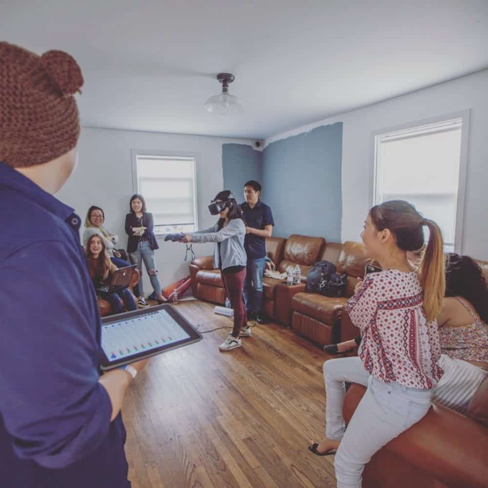 People in a living room with VR user, watching and laughing. Someone holds a tablet.