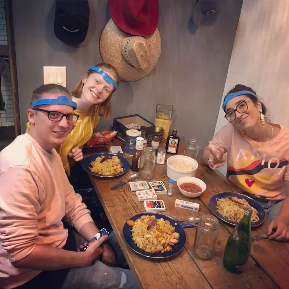 Three people wearing headbands at a table with food and a game, smiling, casual setting.