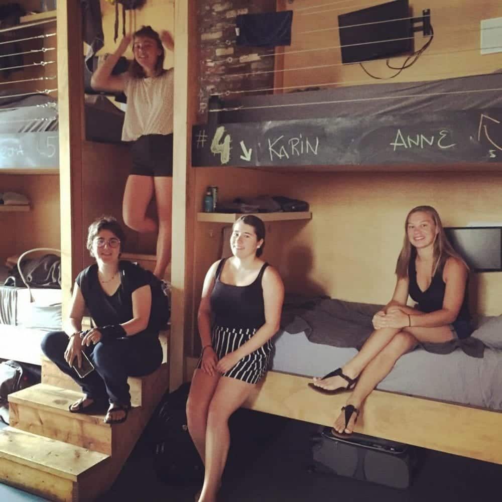 Four young women in a hostel. One stands, others sit on bunks. Wooden interior.