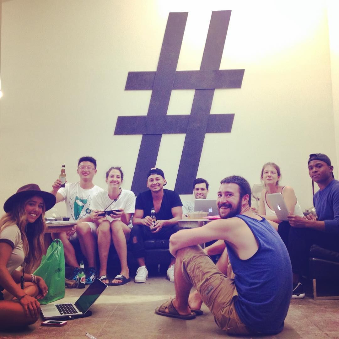 Group of diverse people in a brightly lit room with a large hashtag symbol on the wall, some working on laptops.