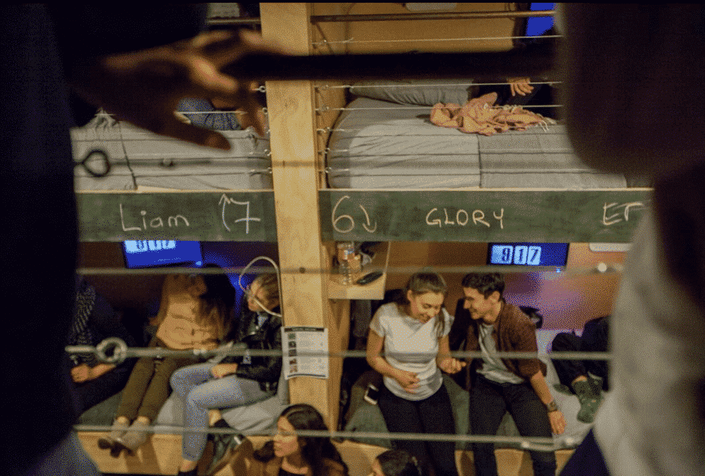 Bunk bed dorm room with people, including a couple laughing together. Chalkboard signs read 