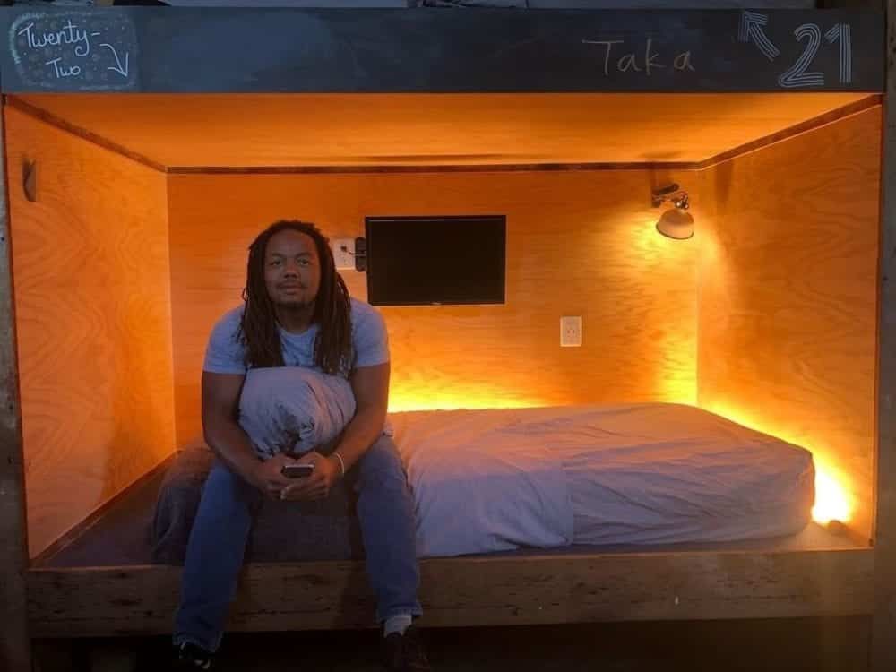 Man seated on bed inside a wood-paneled cubicle; warm lighting; small TV, 