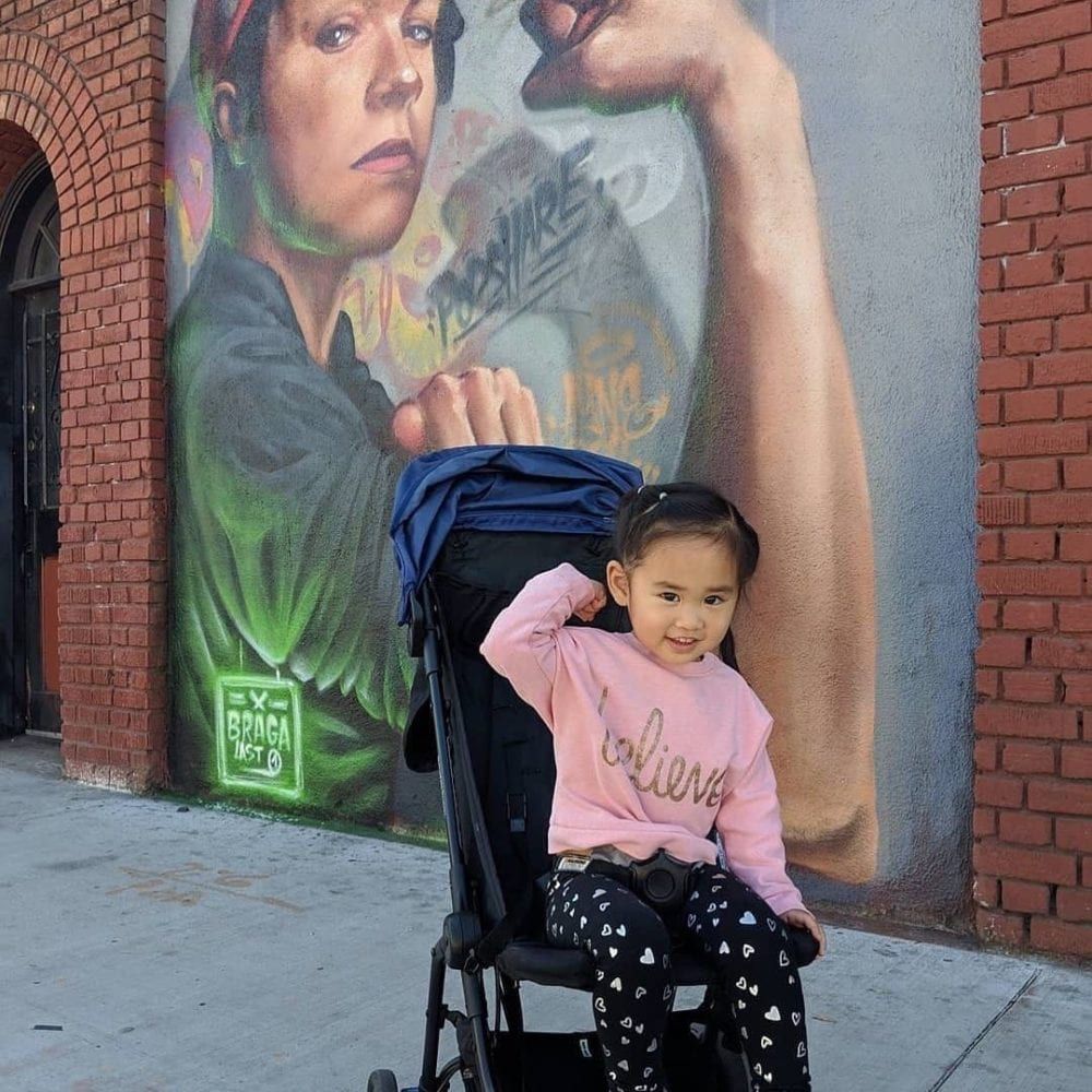 Girl in stroller flexing arm in front of a mural of a woman flexing. Brick building backdrop.