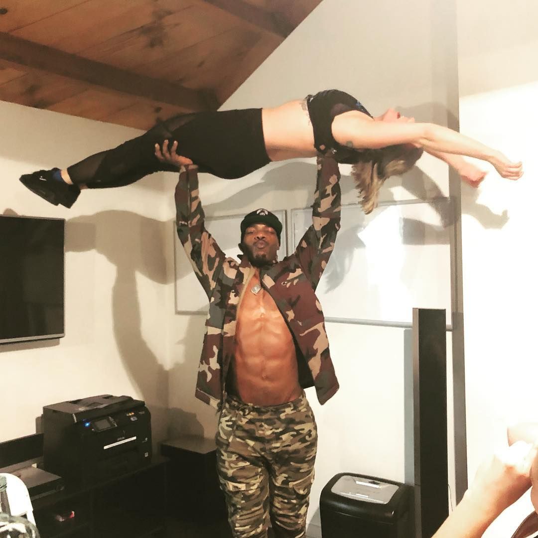 Man holding a woman horizontally above his head. Both are wearing workout clothes in a home setting.